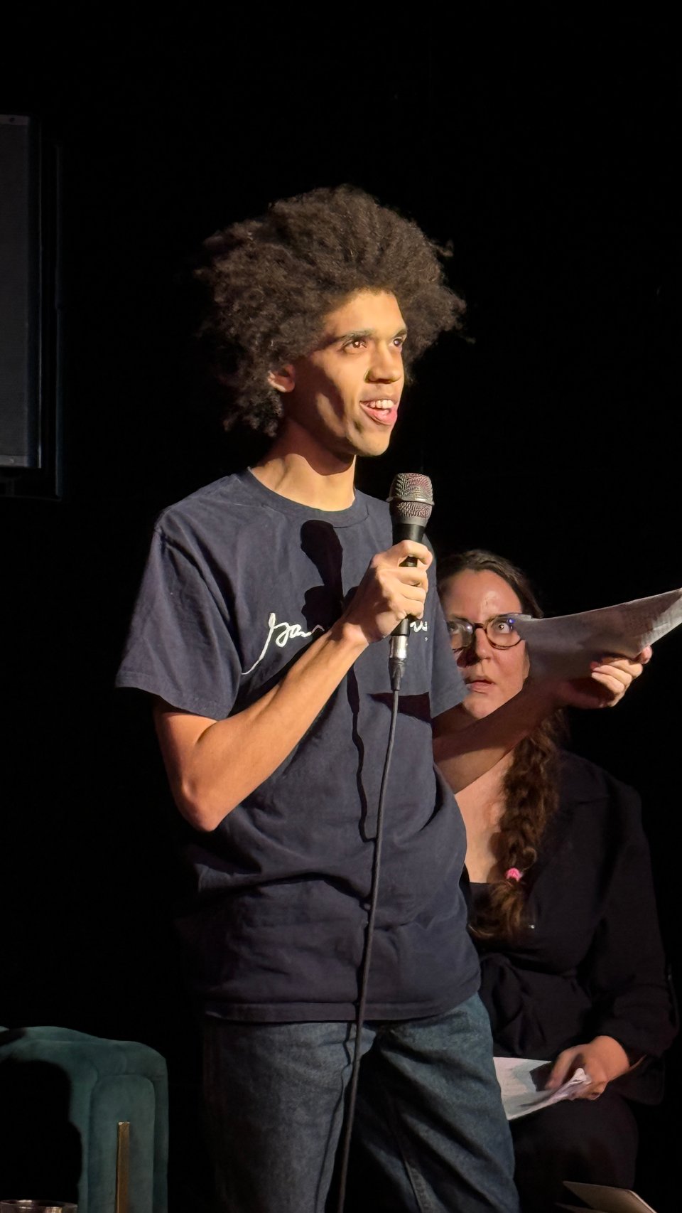 Ciarán Finlayson standing with a microphone, looking at the audience with his manifesto script in hand. Ania Szremski, seated behind him to the left, watches.
