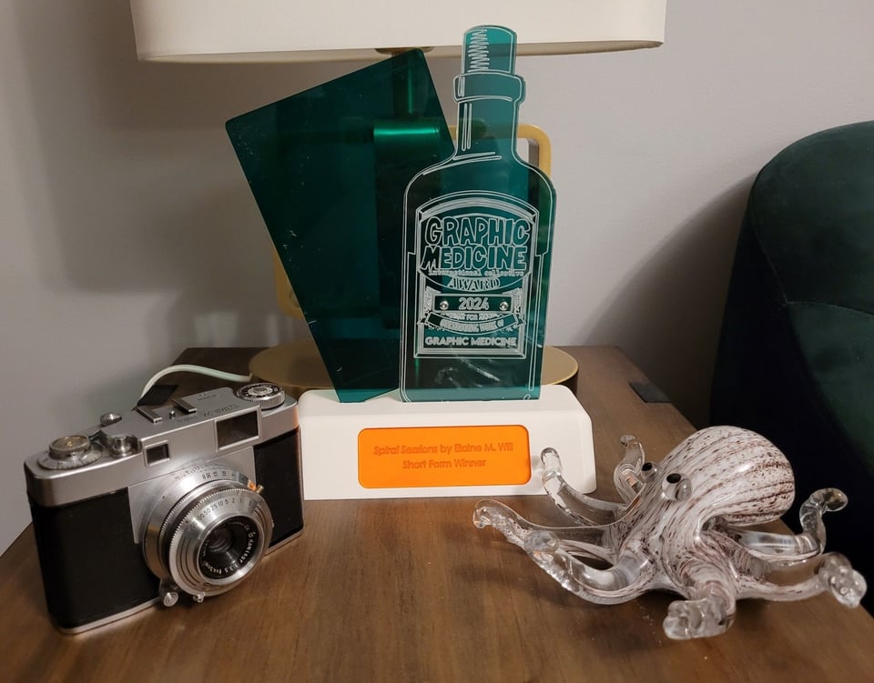 My Graphic Medicine Award trophy for Best Shortform Comic, "Spiral Sessions." The trophy is composed of two flat pieces of green plastic fastened to each other: one an old fashioned medicine bottle reading "Graphic Medicine Award" and the other a tilted comic book page behind the bottle. Both pieces sit on a white plastic base. The trophy is flanked on the right side by a glass octopus, and on the left side by an old broken film camera.