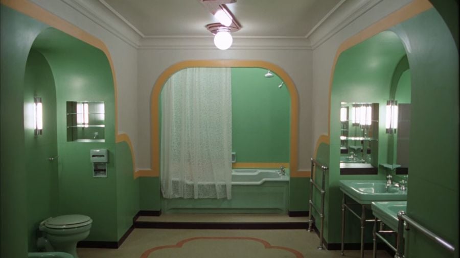 A large bathroom in mint green, with a white ceiling and some decorative flair, and pale yellow trimming. Sinks are on the right, a toilet on the left, and straight ahead is a bathtub with the curtain drawn half across it.