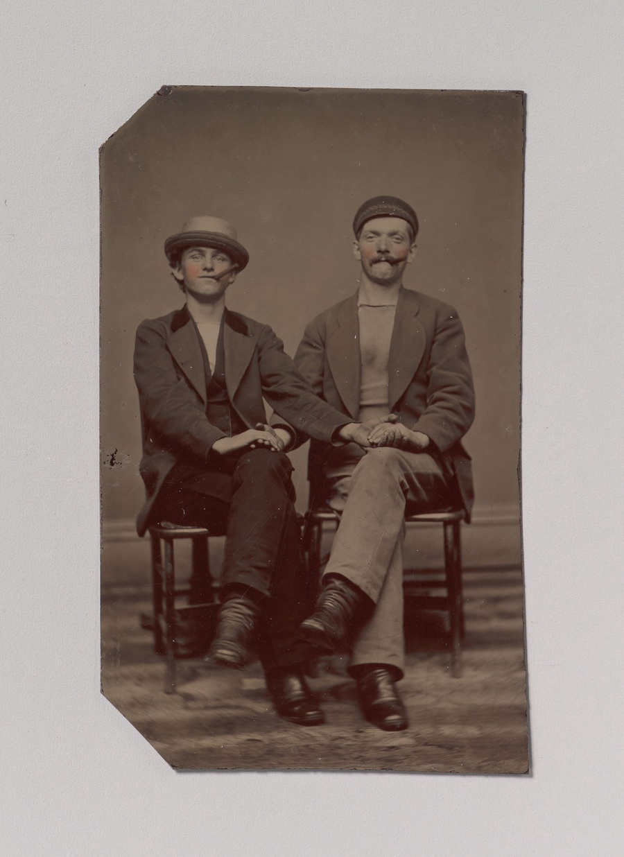 A sepia-toned photograph of two pale-skinned men sitting next to each other on chairs. They are holding hands so that their arms cross into each others laps. They are both holding cigars in their mouths and their cheeks have been hand colored in pink. The man on the left is wearing a straw hat and a suit. The man on the right is wearing a cap and a blazer over a crewneck shirt.
