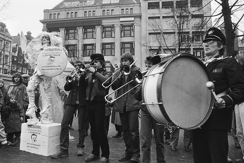 Black and white photo of a 1983 union protest against benefit cuts in Amsterdam. There is a crowd listening to a performance by two people with trumpets, two with trombones, and one with a drum.