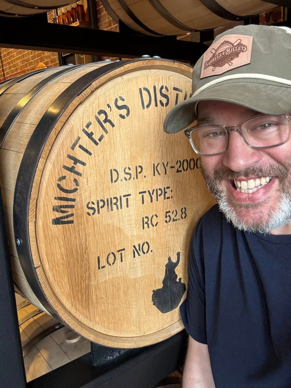 a bearded neil kleid wearing a green barrels and billets hat next to a barrell of michter's bourbon