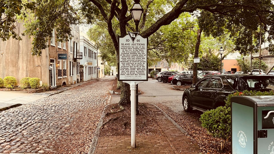 A historical marker in Charleston, SC noting the history of that space as a slave marketplace. It is centered in the photo, alongside a cobblestone lane on one side and parked cars and a solar powered compacting trash can on the other. The sign appears to stand in the middle between the past it retells and the present the reader stands in while reading it.