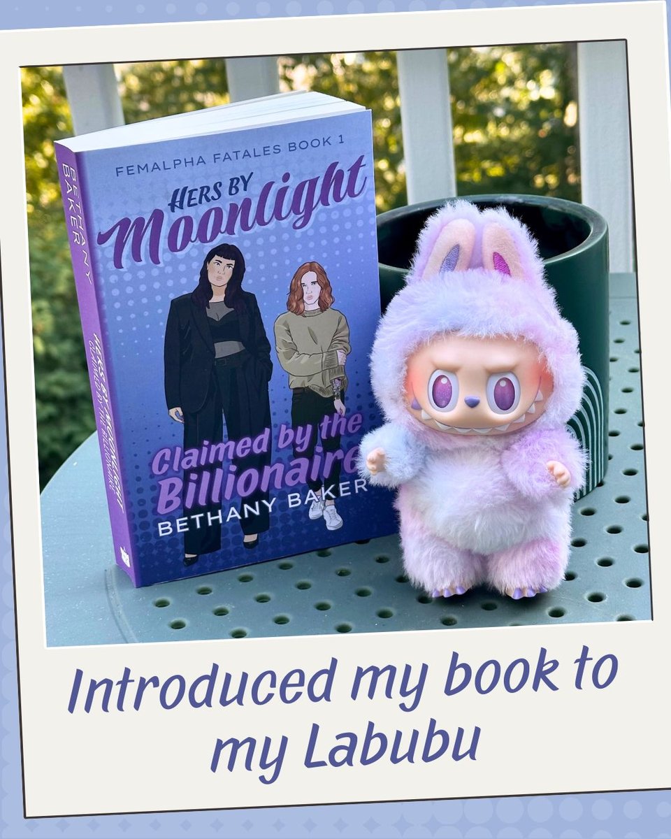 A photograph of the book (cover as previously described) sitting upright on a table next to a purple and pink Labubu doll. The doll has a fuzzy body, bunny-like ears, and a molded vinyl face with big sparkly eyes, a button nose, and a wide toothy grin. It's giving gremlin energy. Text: Introduced m y book to my labubu