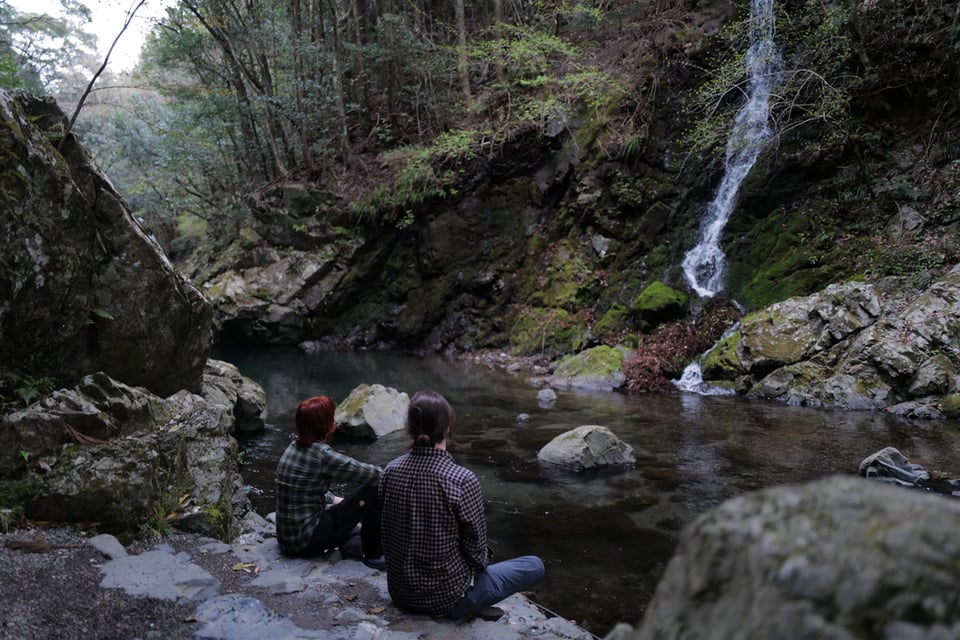 Two people sitting quietly on rocks beside a tranquil moss-edged pool, gazing at a slender waterfall cascading through a forested gorge