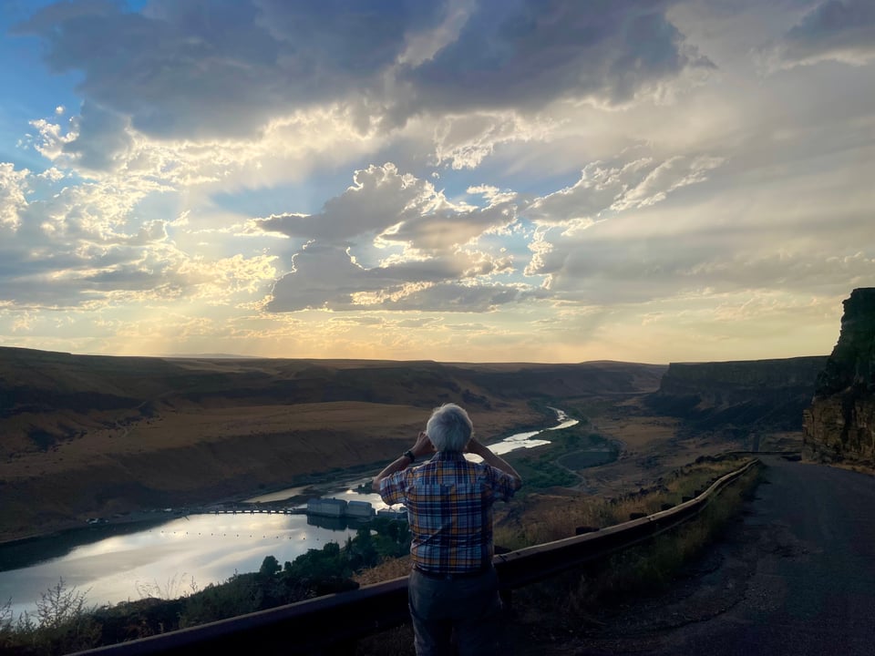 a man with silver hair looks out onto a river and a sunset and takes a photo