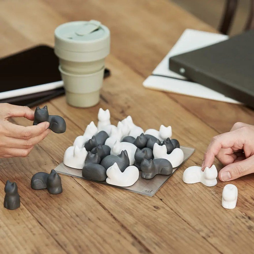 A photo of the game, showing perhaps a dozen of the cat-shaped game pieces nestled together on a small grid board. Half the cats are black, and half are white; some are shaped in an L shape and some a straight. The cats are posed as if they were lying down crouched on their bellies, their heads and backs pointing up. They are fairly abstract shapes; you can see the ears and paws and body and head, but no eyes or mouth or nose