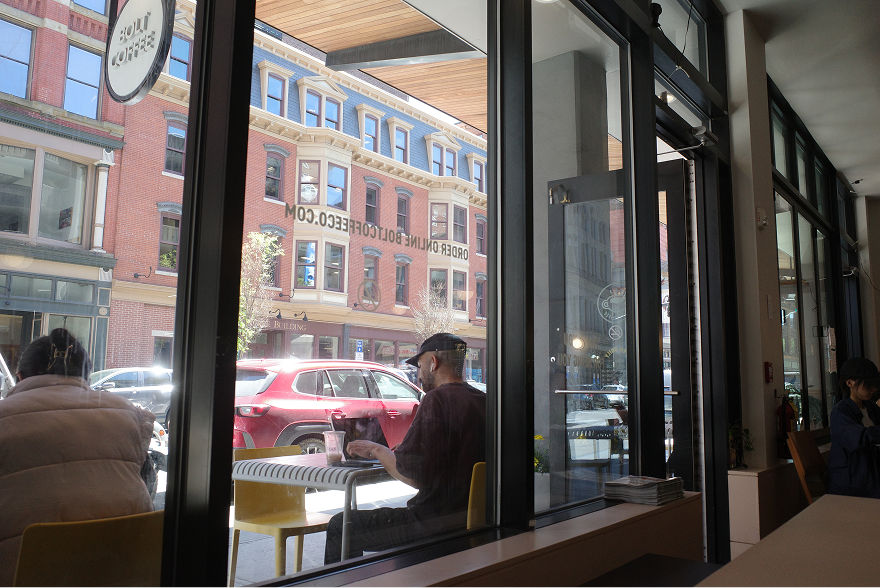View from inside a coffee shop looking out through tall windows onto the street, with people sitting at a table by the window.