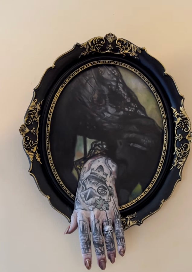 An oil painting of a human arm possibly laying on an armchair handle, wearing a black sheer lace dress with long sleeves. From the painting, a sculpture of a hand comes out giving a 3D impression to the art. The canvas is oval and framed with black and golden rococo details.