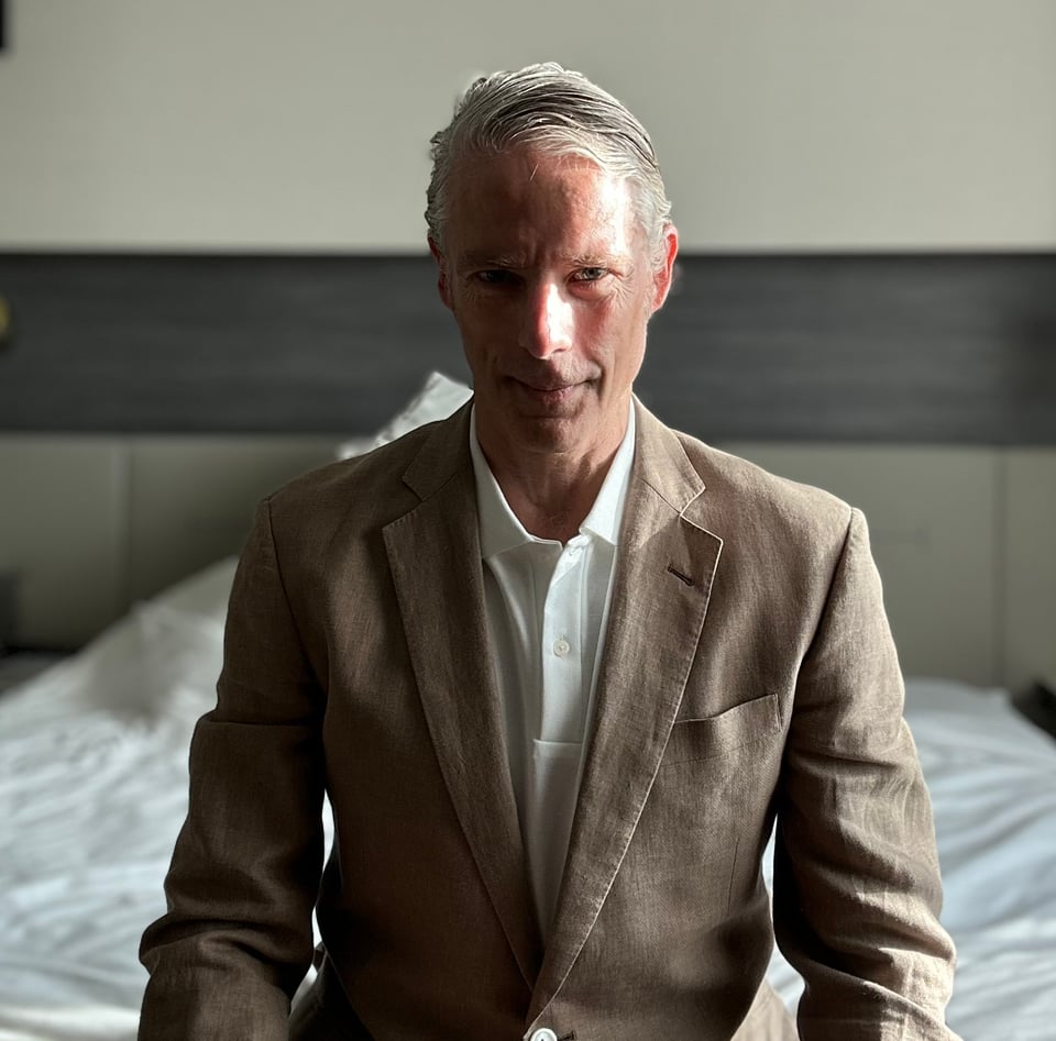 Photo of white man in light brown linen suit & white polo shirt sitting on a hotel bed. Light from the window his left is strong, so his right side is shadowed.