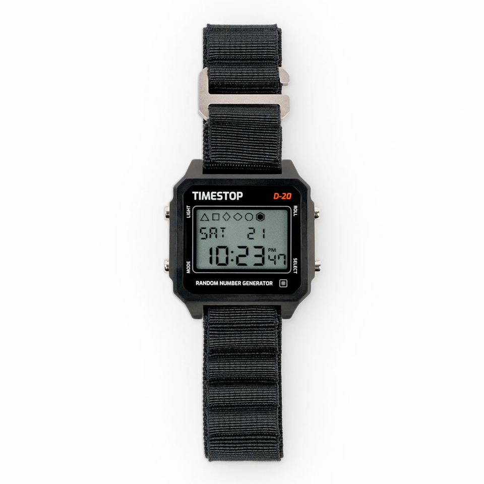 A photo of a black wristwatch against a white background. The brand name at the top of the watch is "TIMESTOP", and the small gray screen has LCDs showing the time of 10:23 PM, on Saturday the 21st. At the top of the screen are six geometric shapes: a triangle, a square, a diamond, a wider diamond, a circle, and a hexagon. The shapes are all hollow, except for the hexagon, the center of which is filled in black. At the bottom of the watch case is printed in white the words "random number generator".