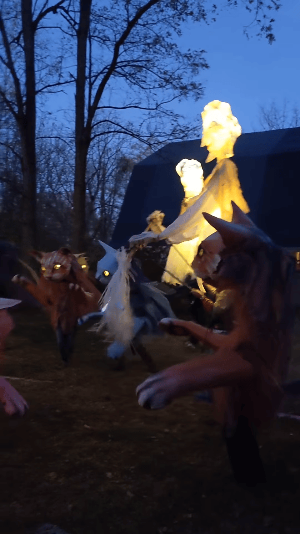 A still image from a group of people with giant cat puppets on their shoulders, and even larger glowing human puppet figures.