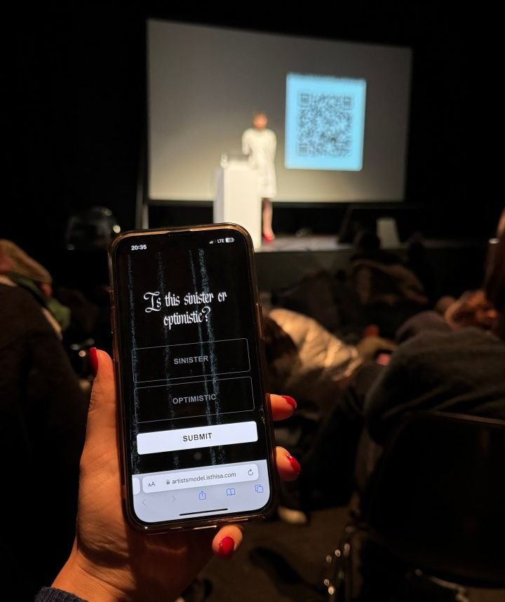 A hand holding a phone in the foreground, and Sarah in the background on stage. On the phone screen is an interface for voting, that asks, "Is this sinister or optimistic?" with Sinster and Optimistic beneath it as voting options