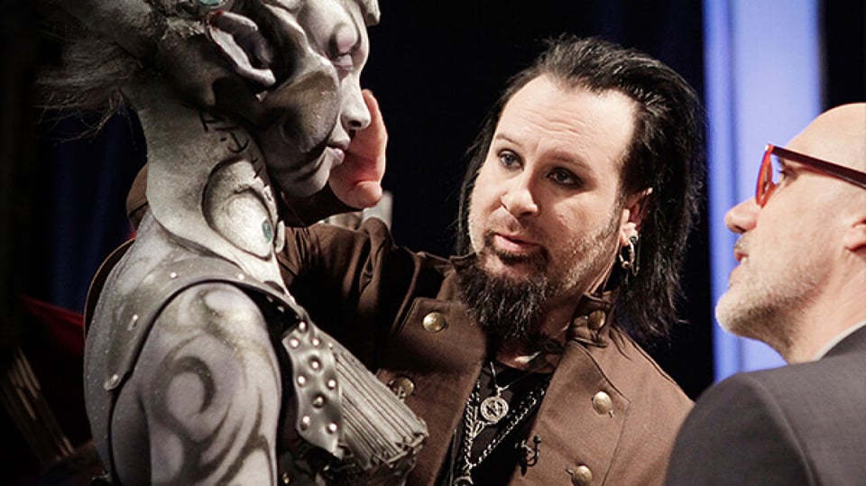 Two people inspect a third person who's in metallic makeup