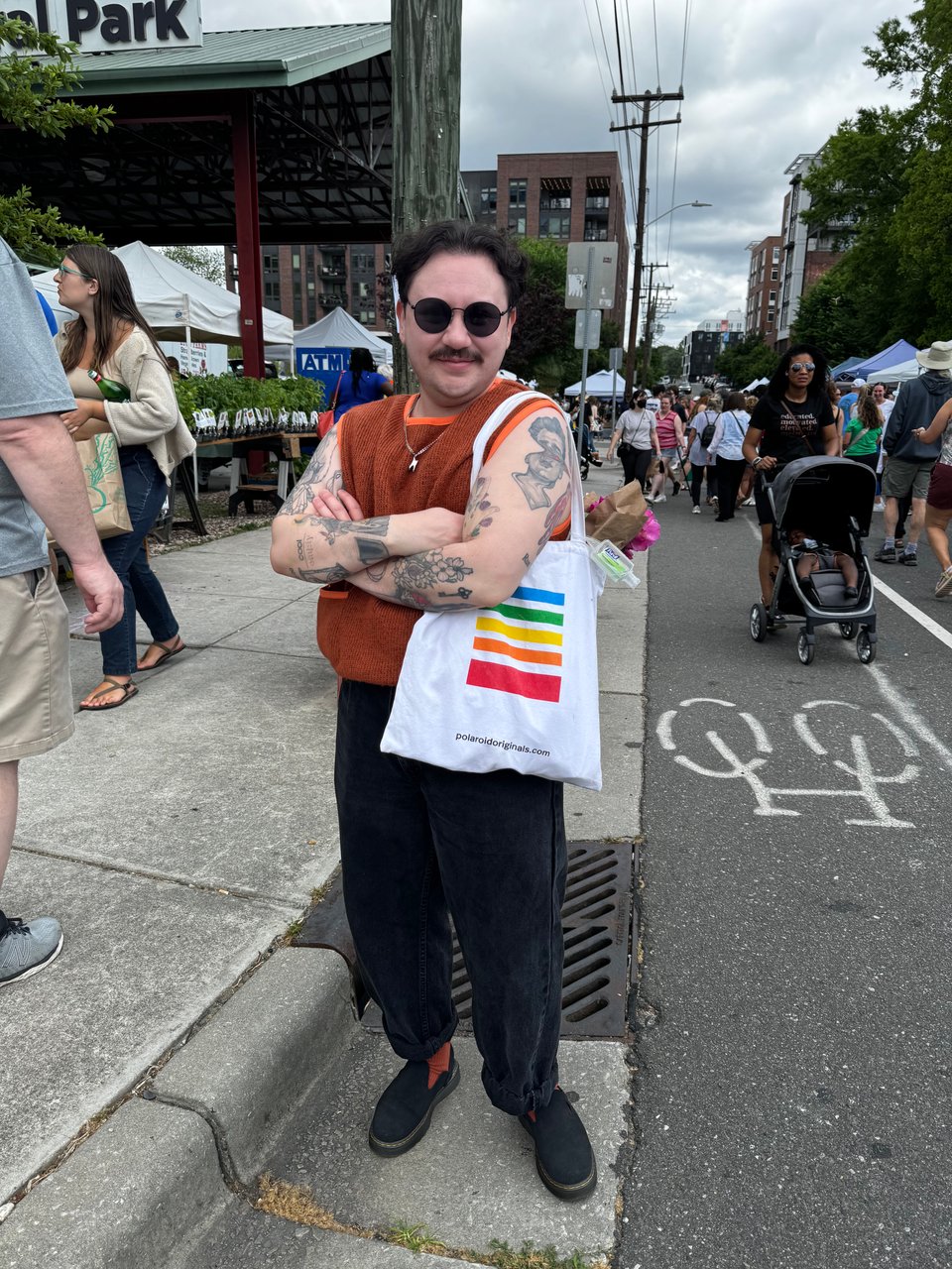 von at the farmers market - he has short curly brown hair, round sunglasses, an orange tank top, black jeans, orange socks, black mules, and a polaroid themed tote bag