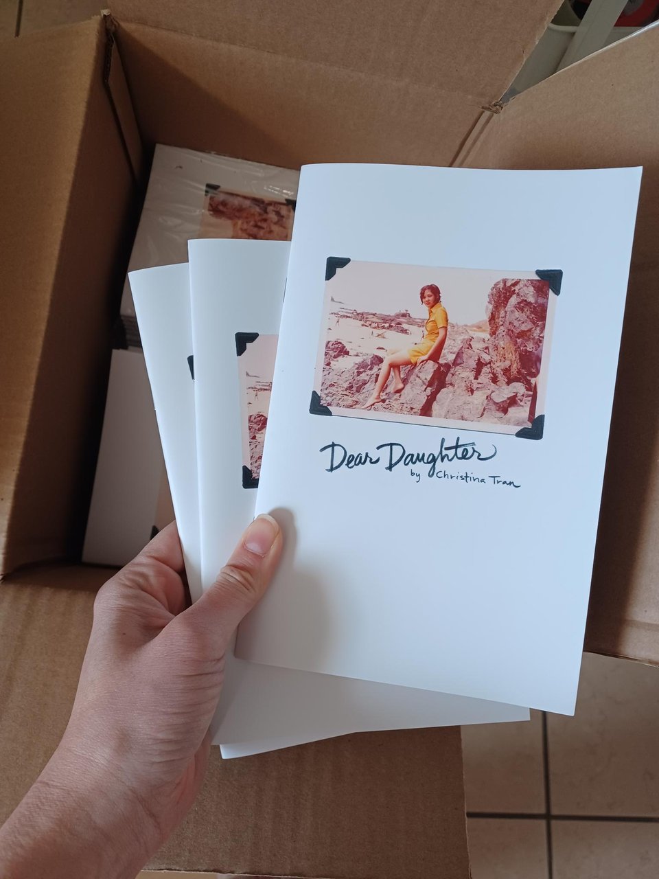 hand holding copies of the "dear daughter" zine against an open cardboard box full of stacked zines