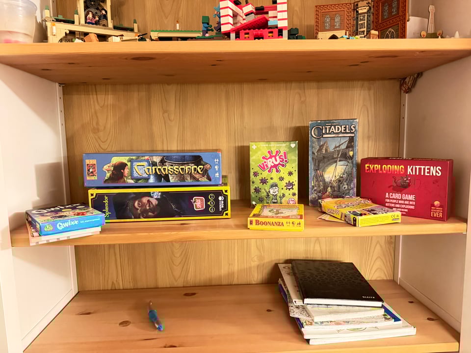 Three mostly empty bookshelves. Top has some legos, middle has eight board games and the bottom has some books and a pen.