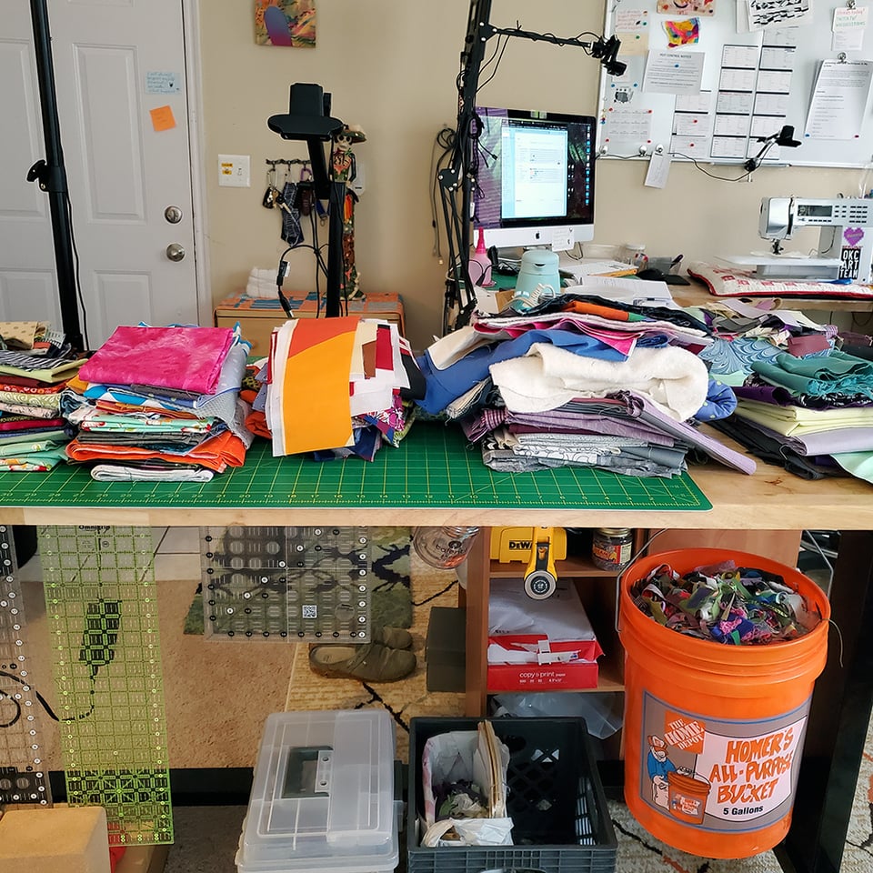 Photo of Sarah's work table with piles of fabric filling the surface. Below the table are Sarah's quilting tools and a bucket of very small scraps.