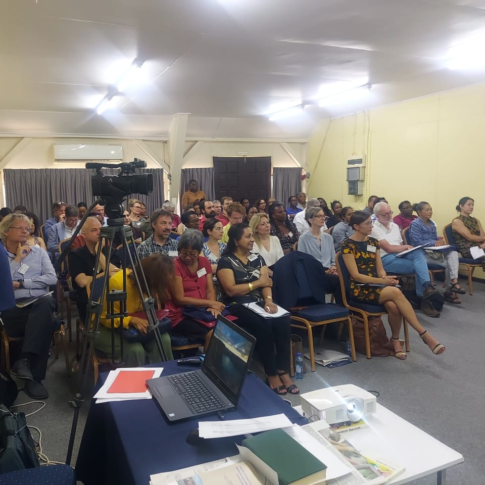 A conference room full with people seated. All seats are taken and there is even a person standing in the back to listen to the speaker.