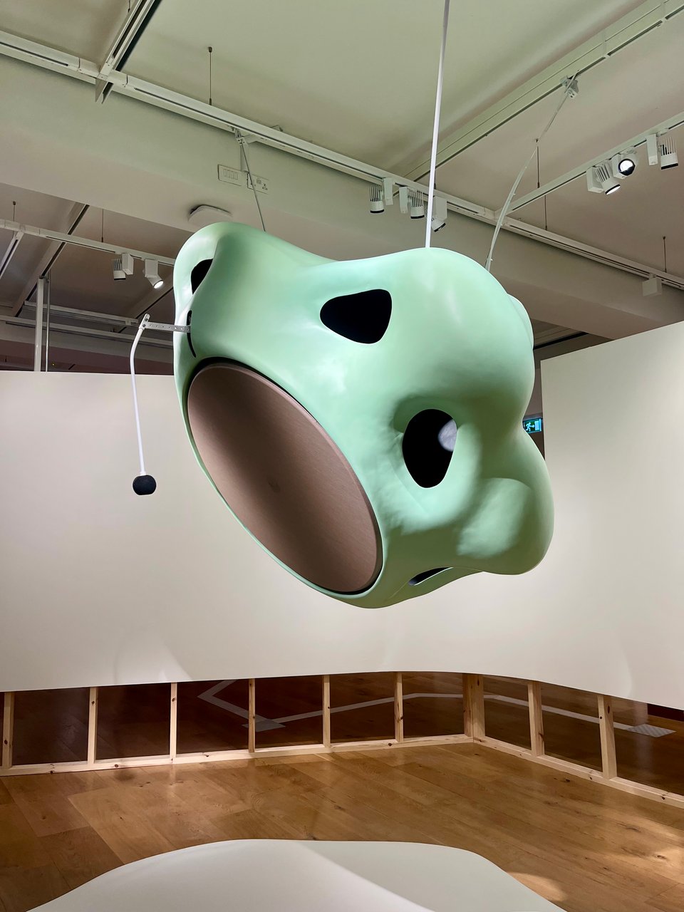 A large green sculpture suspended from the ceilings. It looks like multiple noses stuck together in a circle, with a smooth drum skin under the nostrils. A drumstick on a mechanical arm hangs ready to strike the drum.
