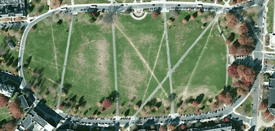An aerial view of a large lawn surrounded on all sides by university buildings. Both paved paths and paths made by walking cross cross over the lawn.