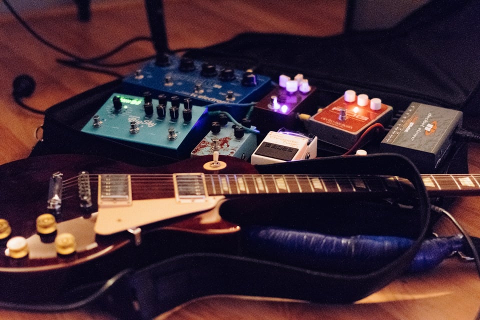 A guitar, on the floor, in front of an array of effects pedals. Photo by Jackie Alexander on Unsplash.