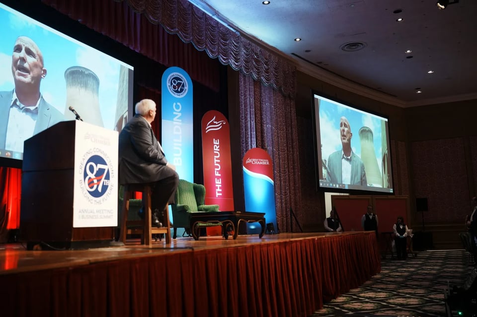 West Virginia Gov. Jim Justice on stage seated, looking at a bald man on a screen as part of a live video call, Jay Powell, in front of an active smoke stack.