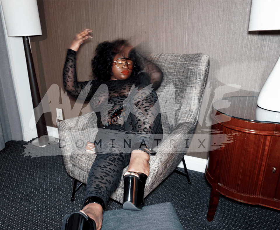 Image of our resident domme, Miss Ava Kane, reclining in an easy chair with leopard print sheer bodysuit and tall black platform heels. She is a larger, confident Black femme domme with big hair and tinted black aviators, with bright red lipstick.