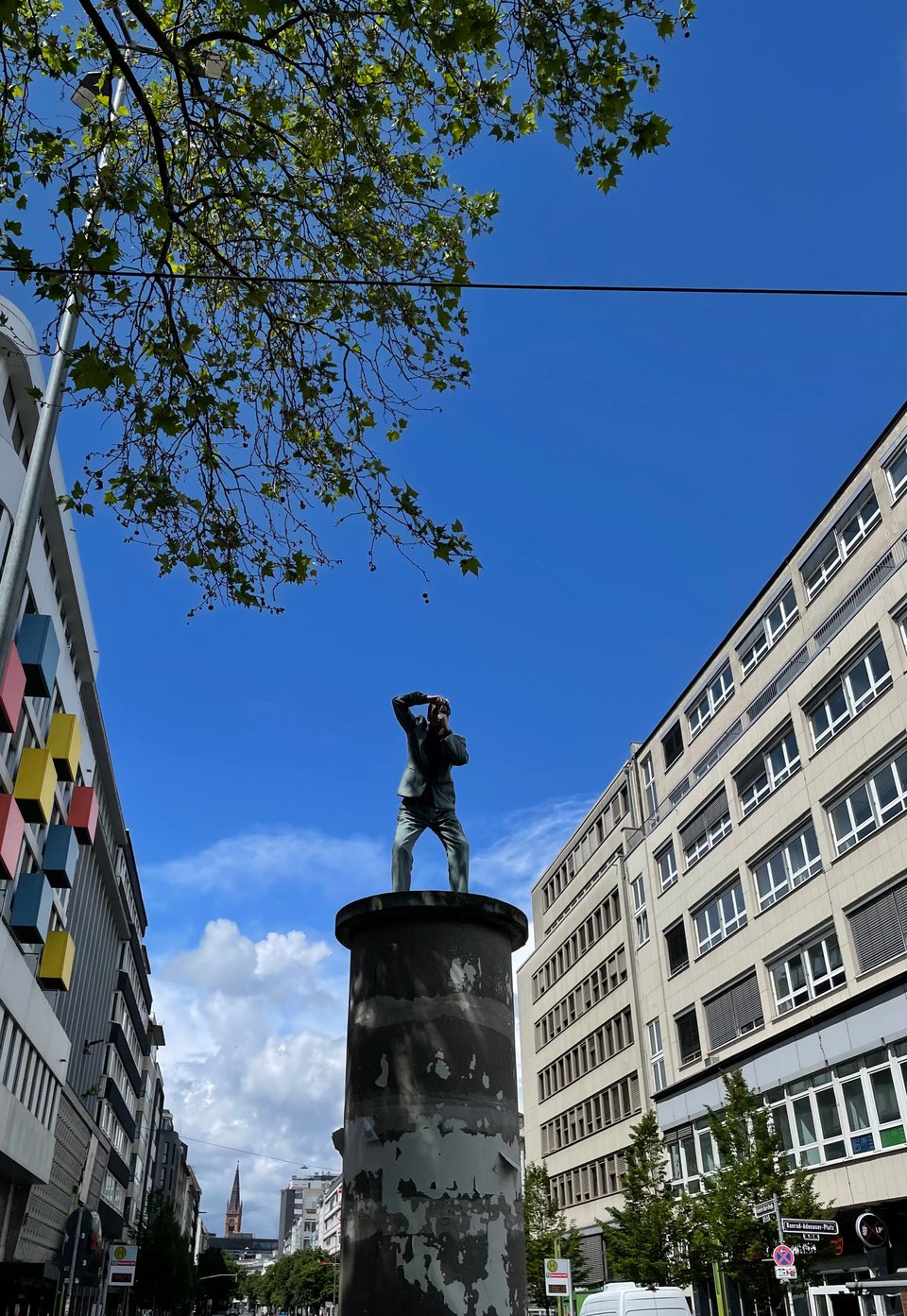 Picture of the sculpture 'The photographer' by Christoph Pöggeler. The sculpture is on top of around 3 metre high pillar, is centred in the photo with a backdrop of clear blue sky with clouds. This is flanked on both sides by buildings on either side of the street