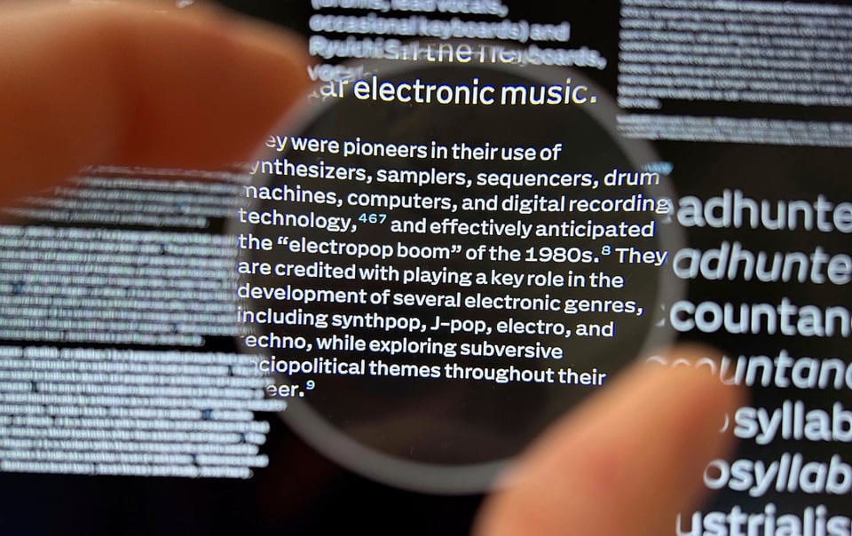 A hand holding a lens in front of a computer display that shows white text on a black background. Despite the low resolution of the screen, all relevant attributes of the letterforms are visible. From the biggest to the smallest shape.
