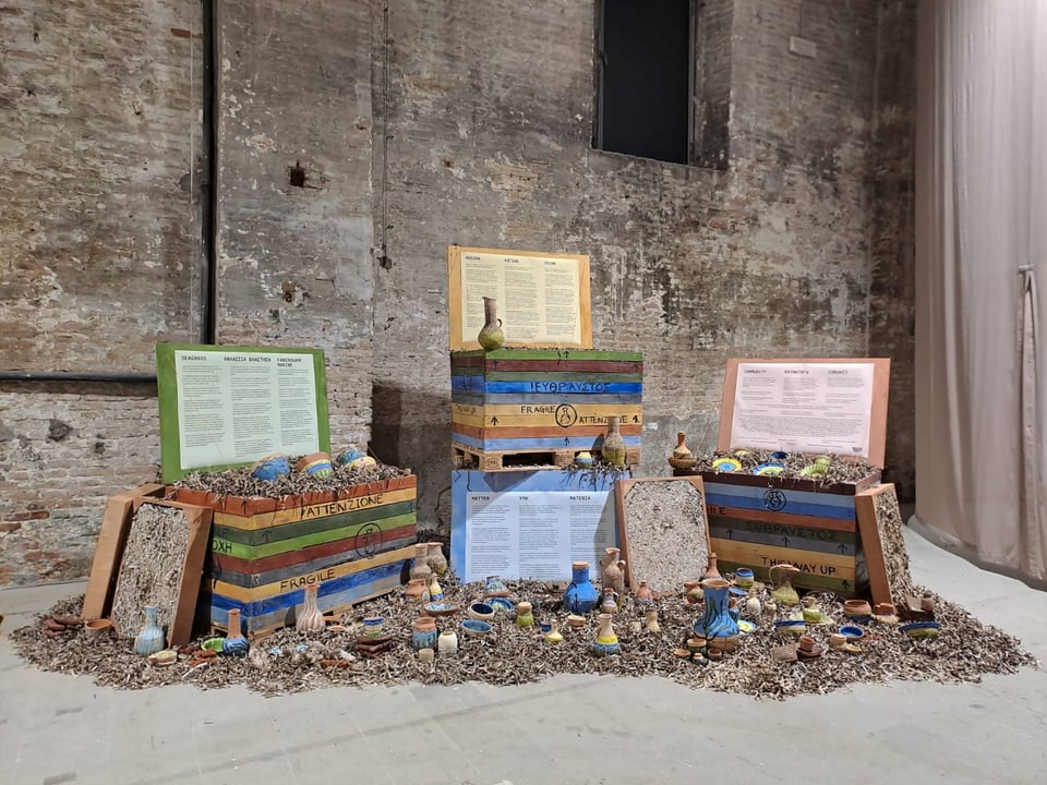 An image of Vessel's installation at the Venice Biennale: four brightly-coloured wooden crates are surrounded by piles of dried seagrass, in which are embedded over a hundred clay vases and other vessels, all painted with seagrass imagery and messages.