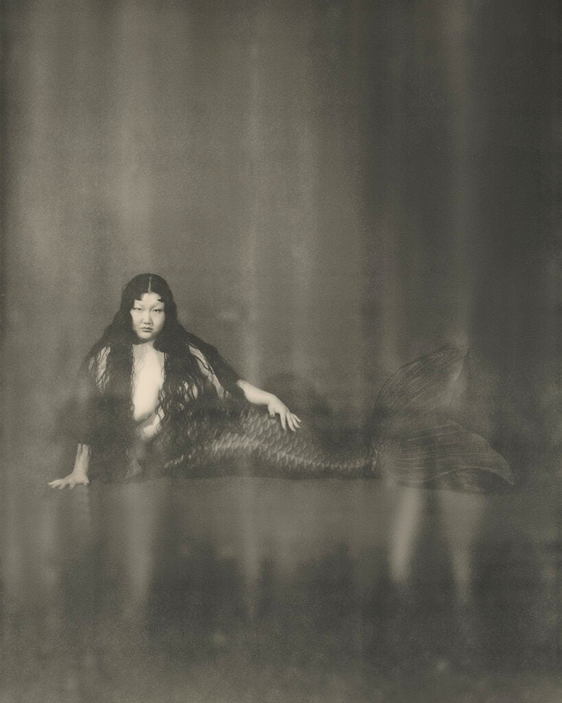 A sepia-toned, soft-focus photograph of a woman posed as a mermaid, reclining on a dark reflective surface with long flowing hair covering her torso and a large scaled tail with fanned fins extending to the right, set against a hazy, mottled backdrop.