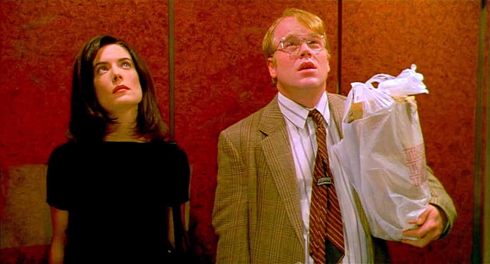 A young white woman with dark hair and a black shirt, standing next to a middle-aged white man with blond hair and a tweed jacket, with his tie turned the wrong way. He's holding a bag of groceries. They're standing in front of a maroon background that looks like the inner wall of an elevator