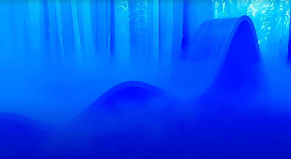 A curved wooden form in immersed in mist and a blue lights in with some projections in the background