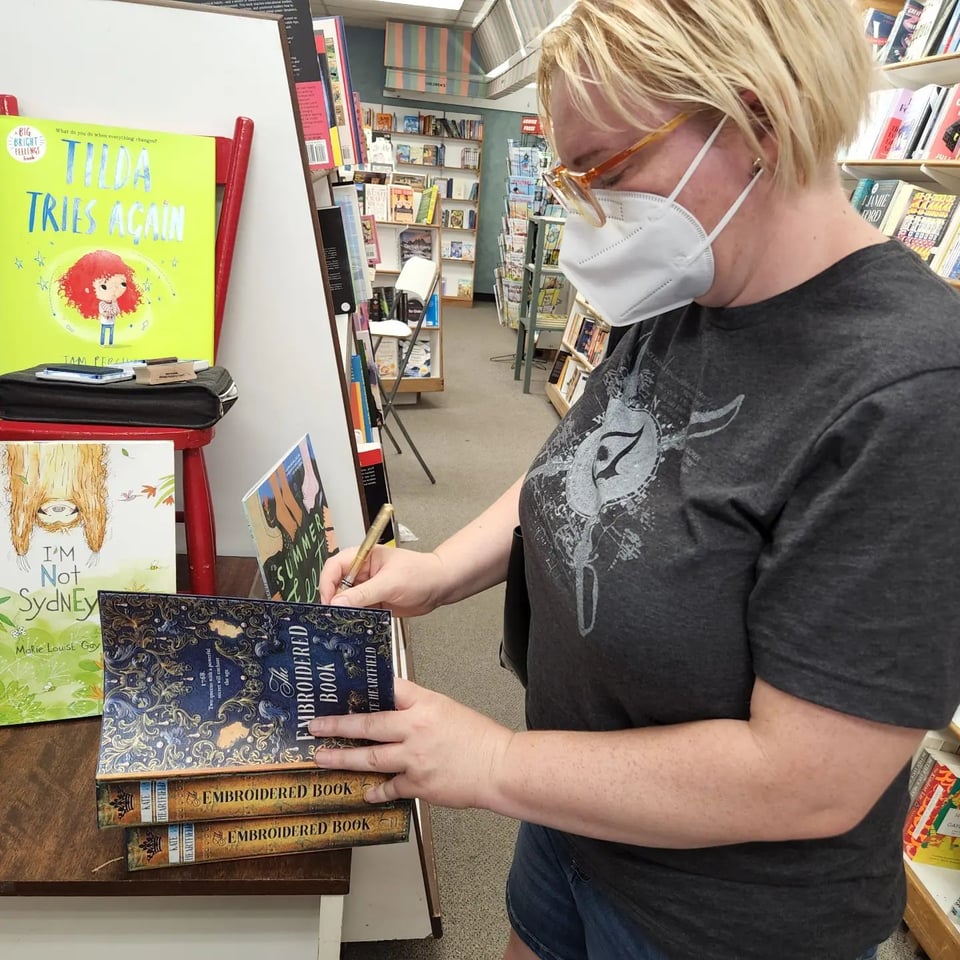 Kate wearing a mask and an Assassin's Creed tshirt and signing copies of The Embroidered Book.