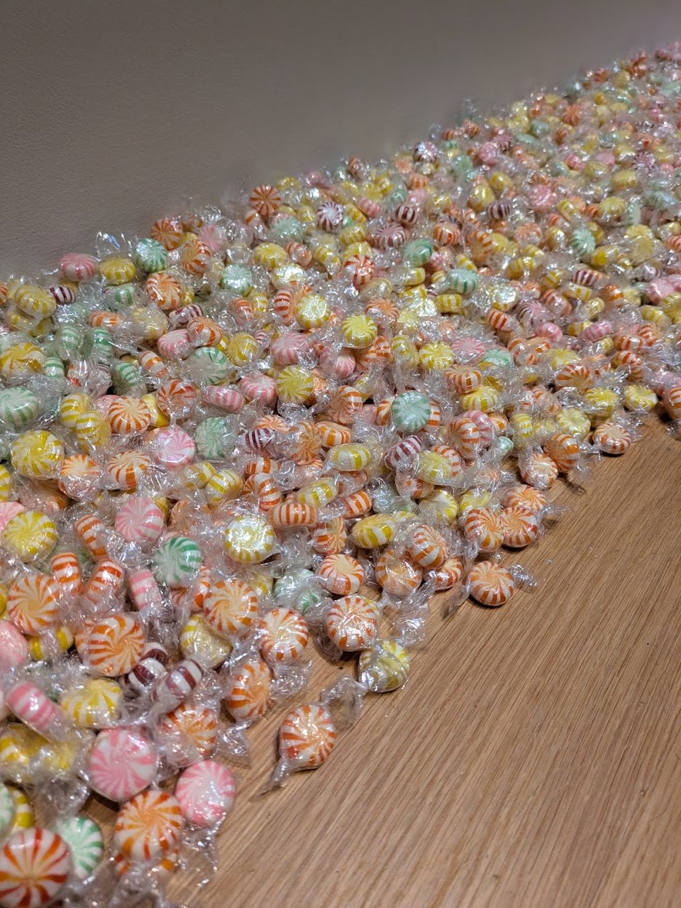 A close-up view of a few hundred striped hard candies in various colors, gathered on a wood floor alongside the wall's edge.