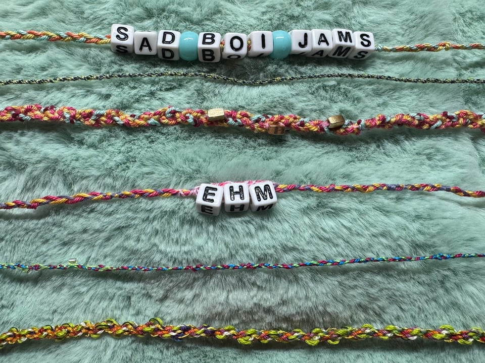 A photograph of colorful friendship bracelets on a green fake fur background. The bracelets say "SAD BOI JAMS" and "EHM".