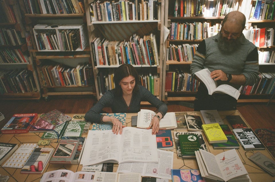 [Two people — one sitting, one standing — leaf through books laid out on a large table. Behind them are shelves and shelves of books.]