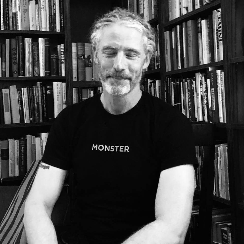 B/W photo of middle-aged white man in black t-shirt that has MONSTER written on it; his eyes are closed