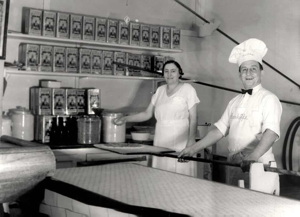 A man in a white chef's uniform with a black bowtie and a big chef's hat holds a pizza peel while a woman next to him, also in all white holds her hand over the peel as if she's about to sprinkle cheese on the pizza.