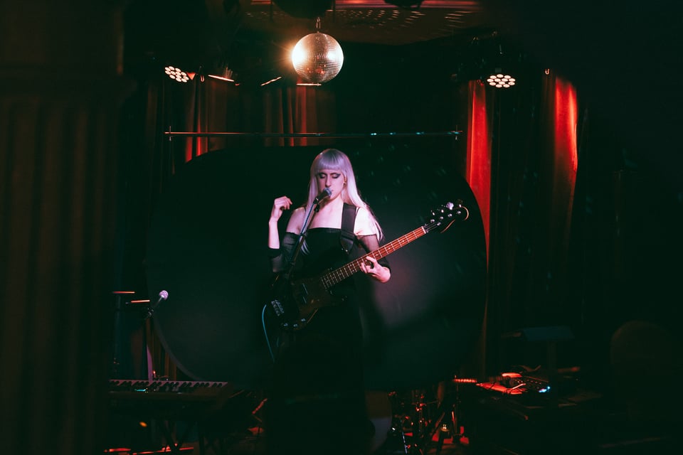 Zir Indigo stands on stage holding the bass, singing with zir eyes shut, beneath a disco ball