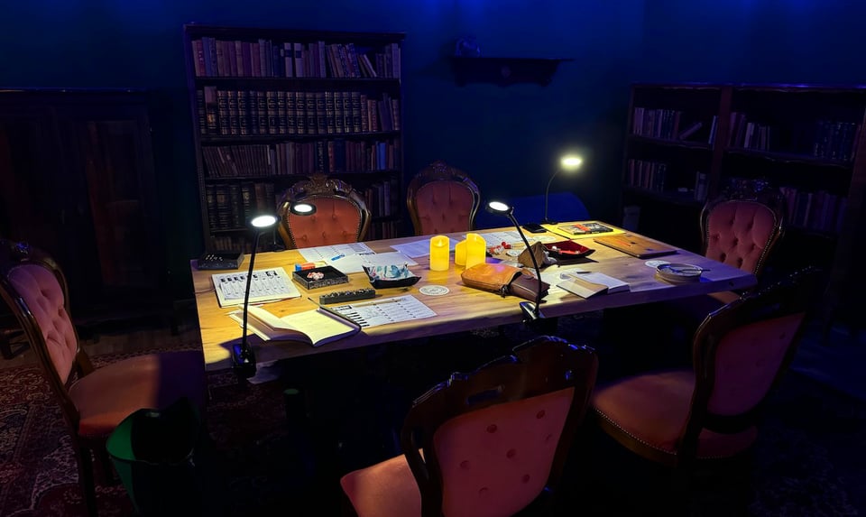 A moodily lit room with a large gaming table surrounded by fancy chairs. Old bookcases line the walls.