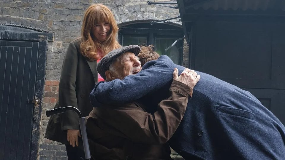Wilfred Mott joyfully hugging The Doctor while Donna looks on with a smile.