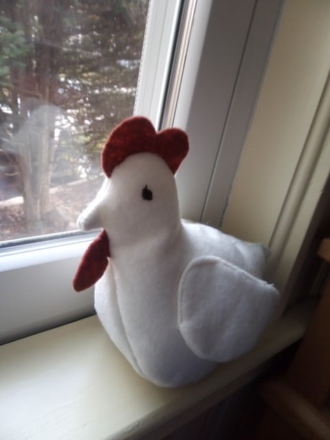 A plush chicken made of red and white felt sits on a windowsill.