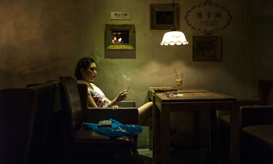 A color photograph of a woman in a shortsleeved white top with a floral design sitting in profile smoking a cigarette at a table with what looks like the dregs of some kind of coffee drink on it. Her bare legs are crossed beneath the table and she looks sad, or at least ill at ease. There are signs on the wall behind her in both Chinese in English, and the light from the hanging white lamp and other sources off camera create undulations of color and shadow on the wall. It evokes melancholy, lost chances, the tyranny of time, the isolation of human beings, the beauty of sadness.