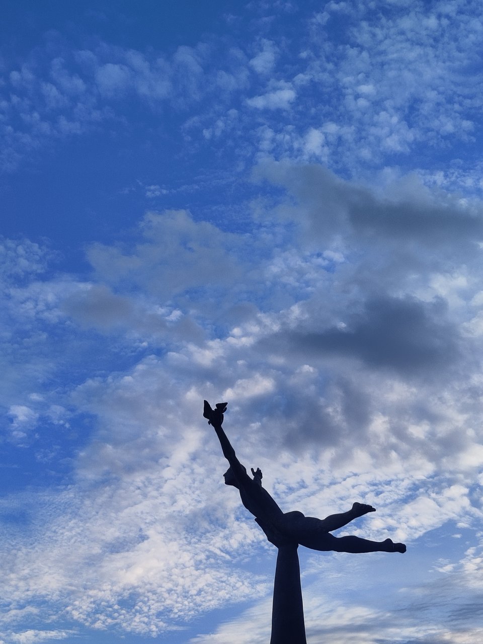 Plastik eines springenden oder fliegenden Mannes vor leicht bewölktem Himmel.