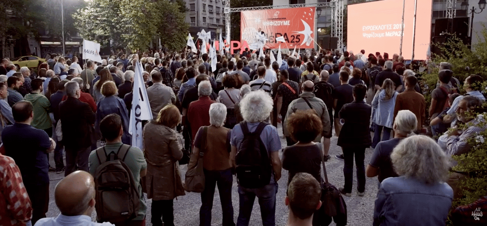 Photograph from the back of a political rally. A crowd of people, some of them holding flags, looks towards a stage on which a politician is speaking. To the right of the stage is a large electronic screen with some Greek text.
