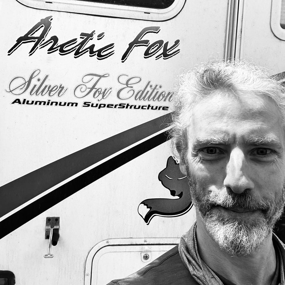 B/W photo of a white man standing in front of an RV that is branded "Arctic Fox: Silver Fox Edition"; since the man has gray/white hair and is conventionally good-looking, this is funny. You can laugh now. It's okay.