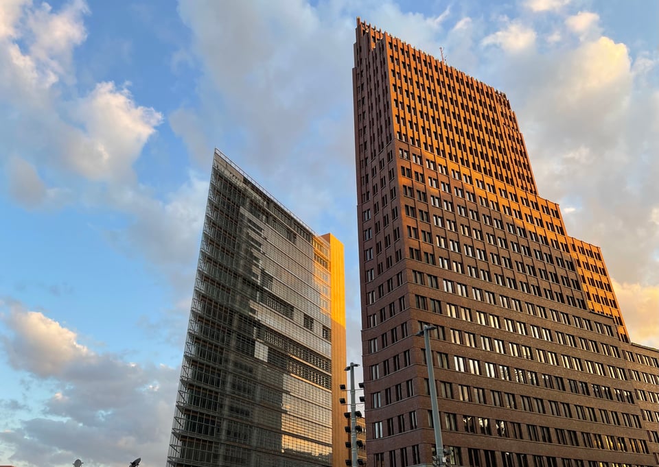 Two buildings stand against the backdrop of late afternoon sky with white, orange and grey clouds. Orange clouds and orange shade on the buildings in general forming by sun going down. The buildings, one brick, another glass, are pointy at one edge. The glass one specially seems like it acts as a knife for the wind
