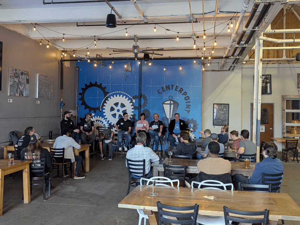 A room with a blue banner saying "Centerpoint" against the back wall, with the Indy Hackers board (most of it) in front and a group of folks in chairs looking at the board and wall from our Open Board Meeting event in April.